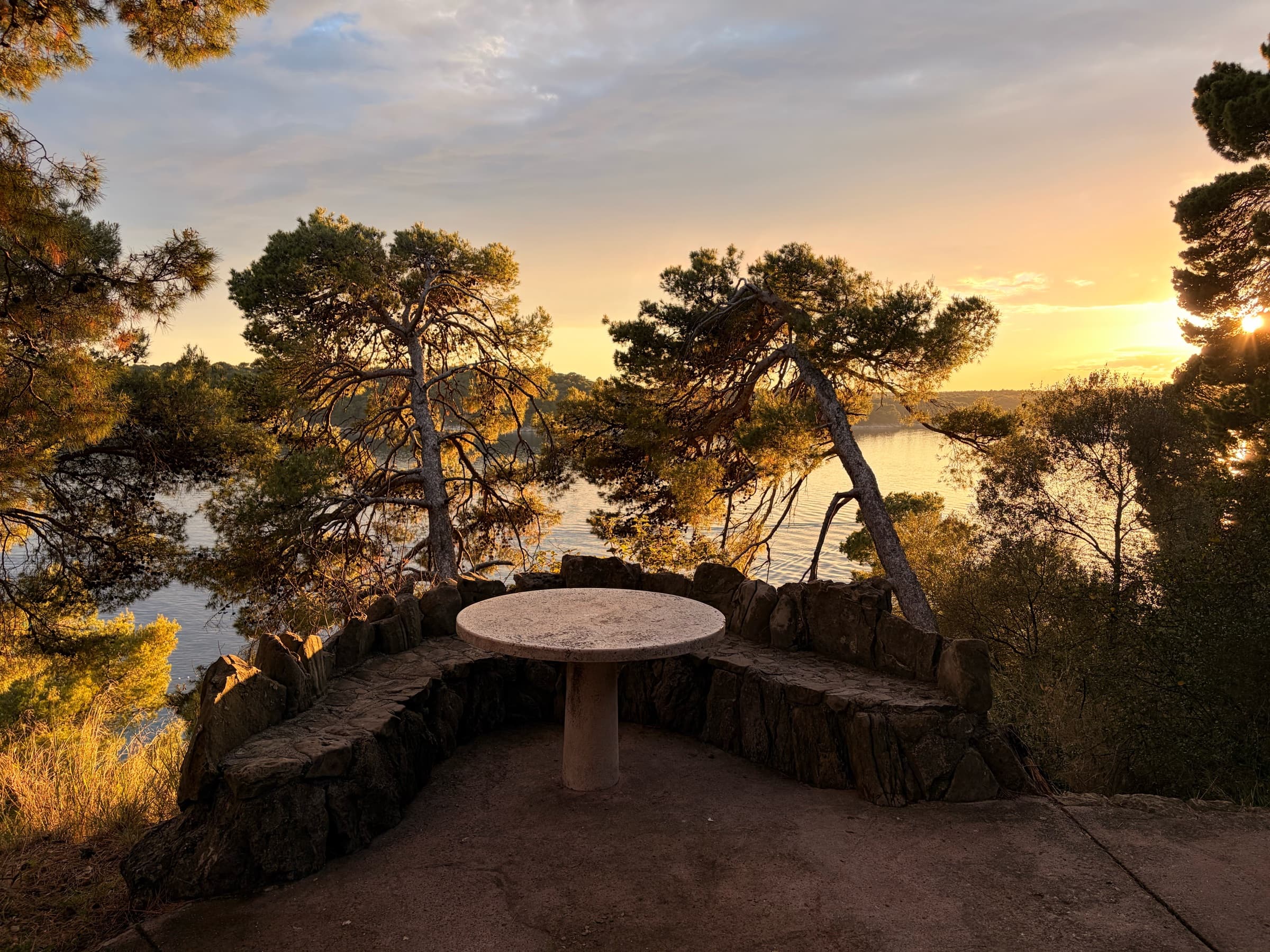 A stone table and bench among pines above the sea in Komrčar Forest Park at sunset.