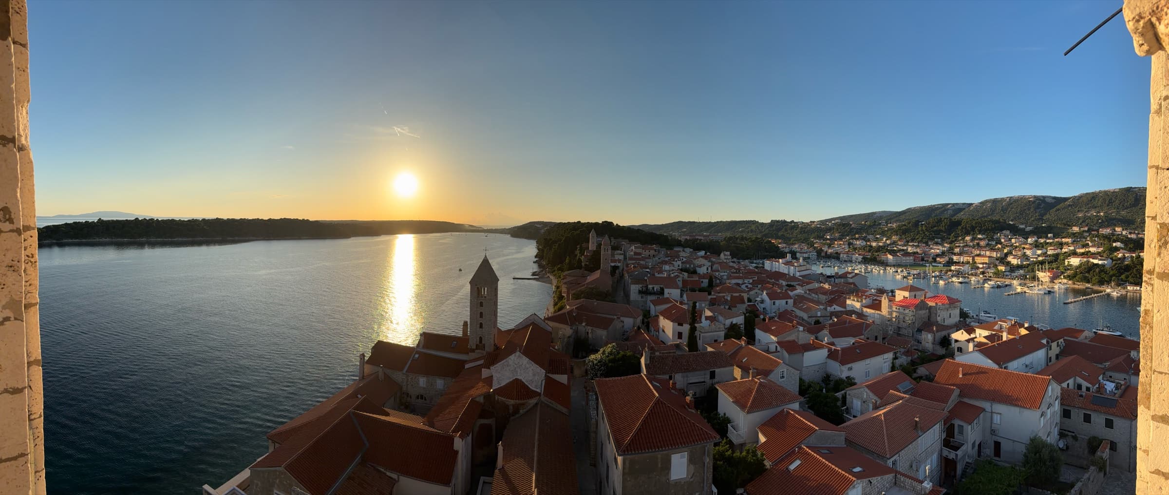 Crveni krovovi, crkveni zvonici, luka i more s katedralnog zvonika pred zalazak.