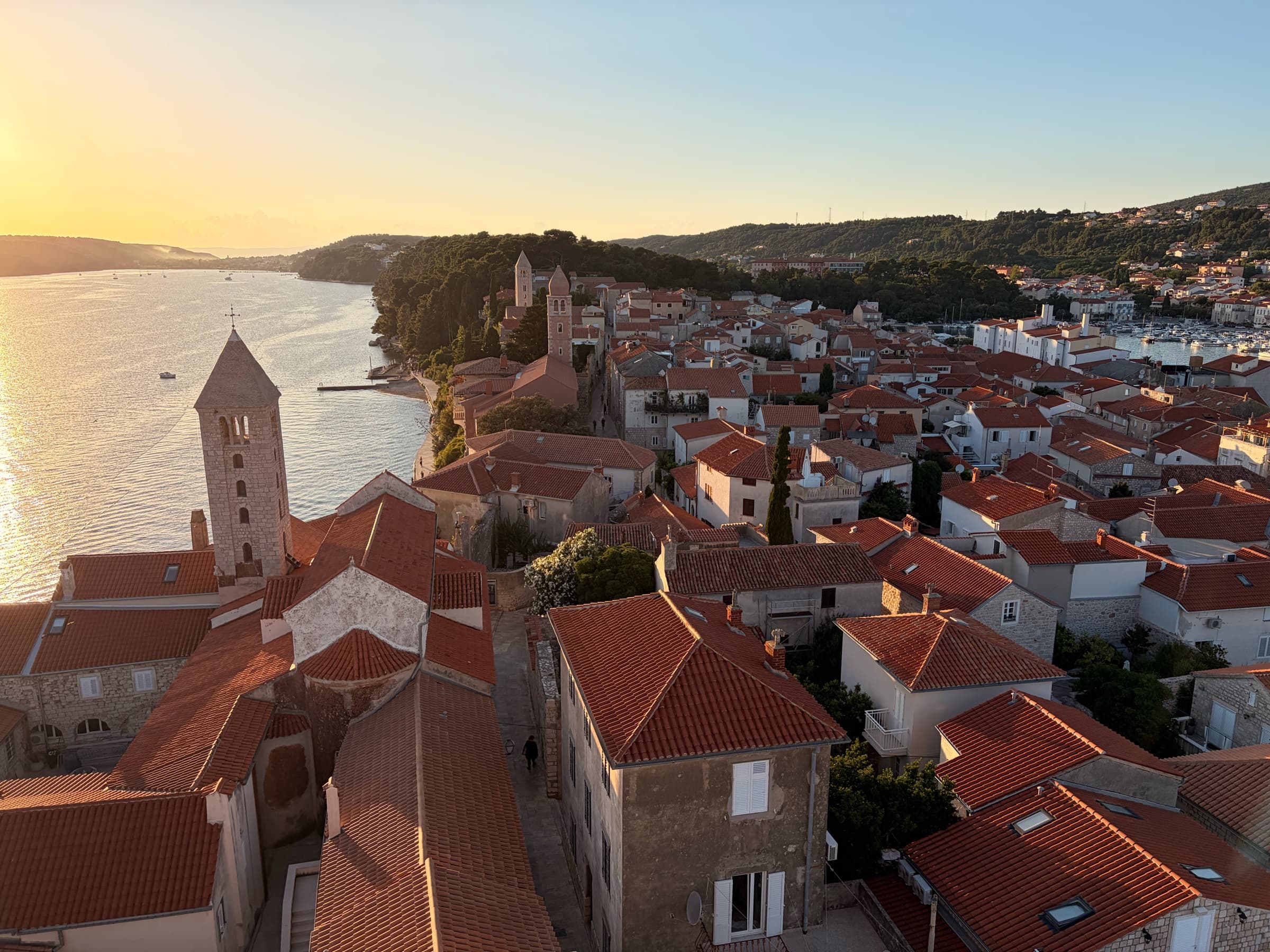 Wide elevated view of Rab's historic rooftops and bell towers in golden light.
