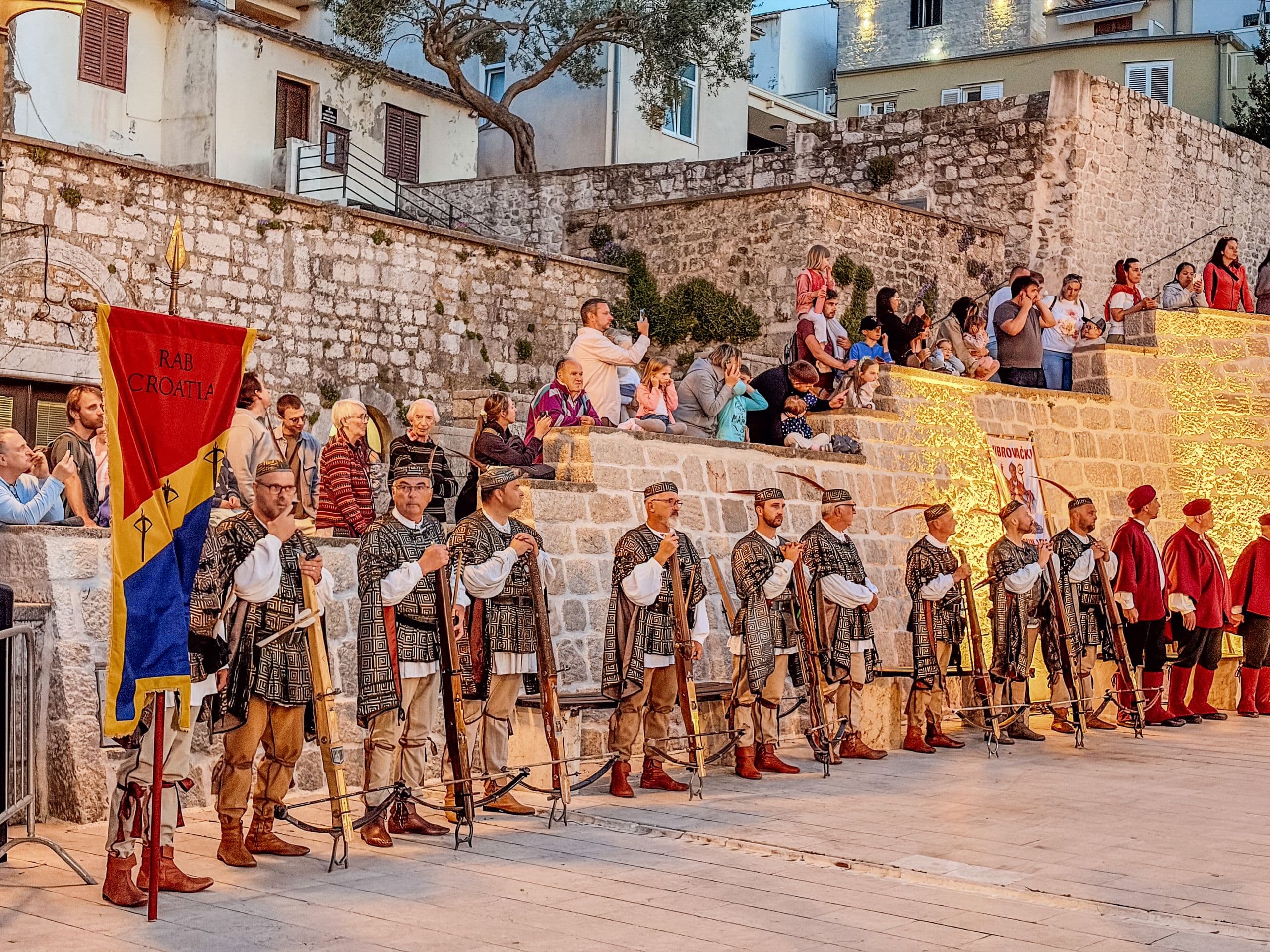 Rab crossbowmen in medieval costume lined up before a seated crowd on the old-town stone steps at dusk during Rapska fjera, a red-and-yellow banner at the left.