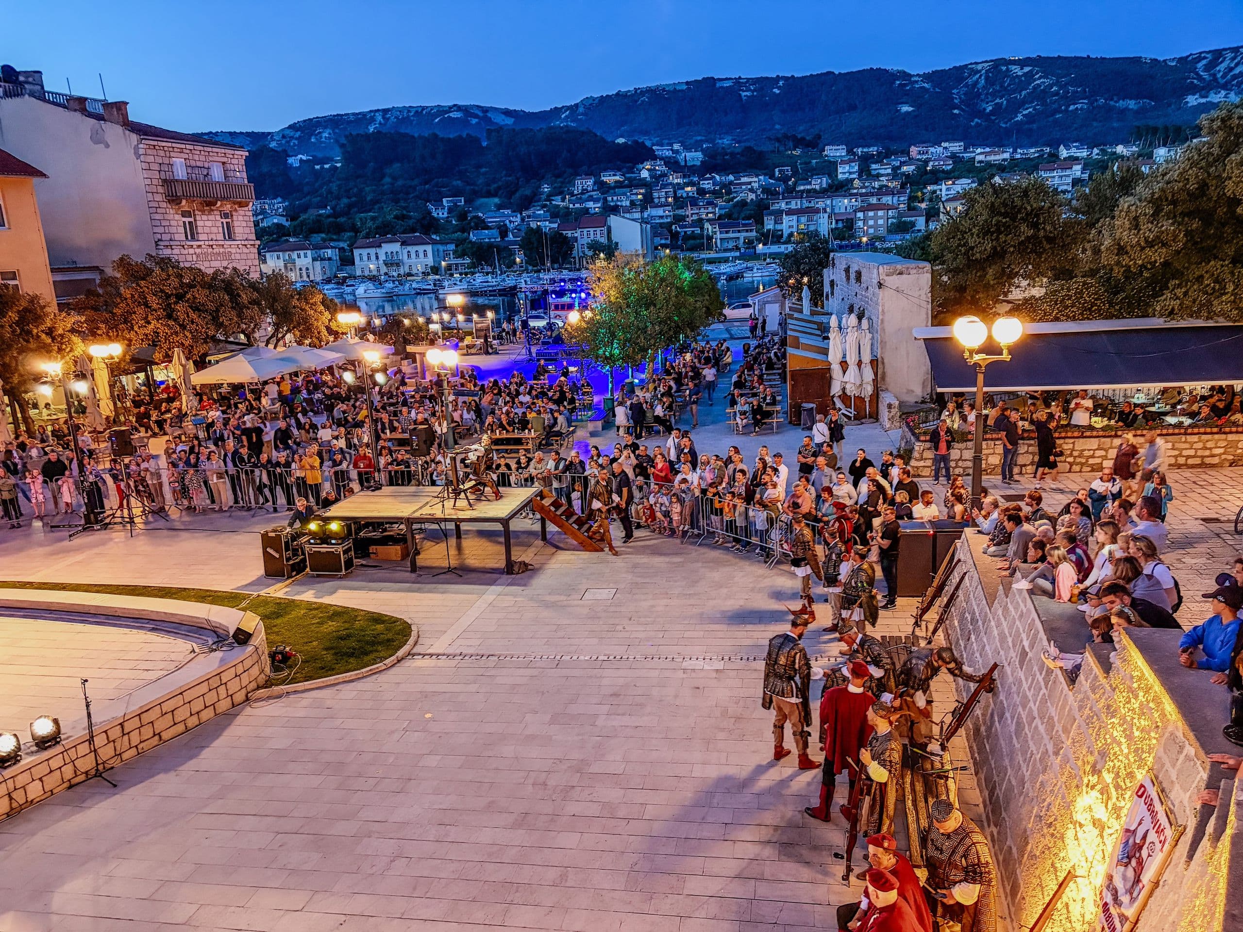 Crowd around an open-air evening stage in Rab old town square, blue stage lights, costumed performers in the foreground, hills behind.