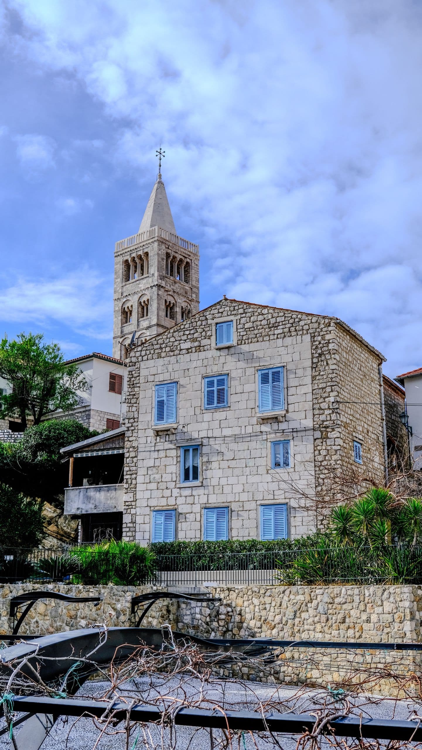 A stone old-town house in Rab, the cathedral bell tower rising behind it under a bright sky.