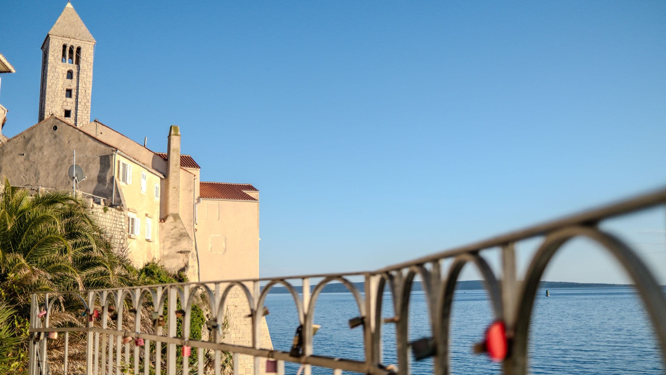 Rab old town rooftops and bell tower above the sea, a love-lock railing in the foreground, bright afternoon light.