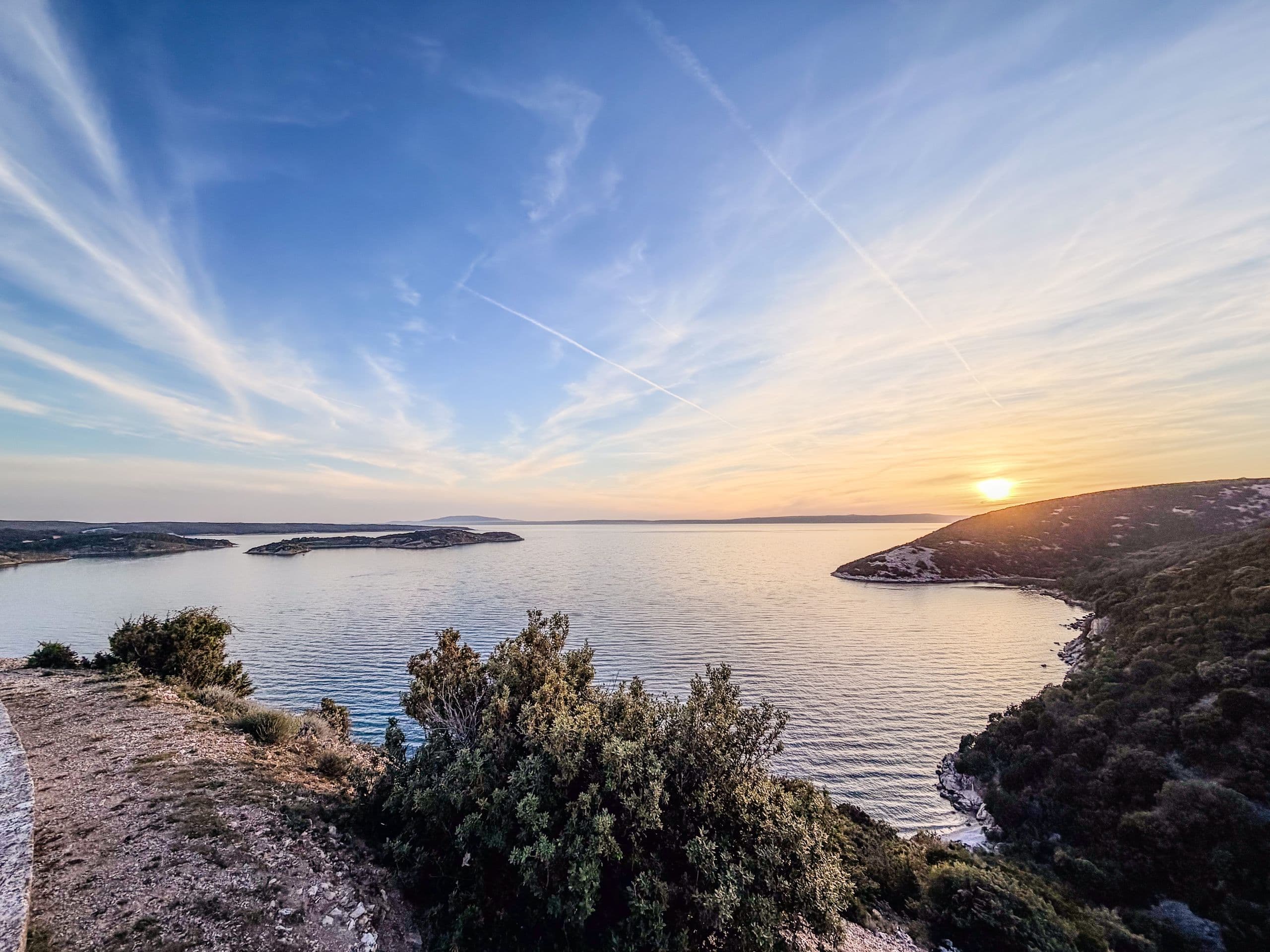 A calm Rab bay at sunset, low hills on both sides, the sun just above the horizon.